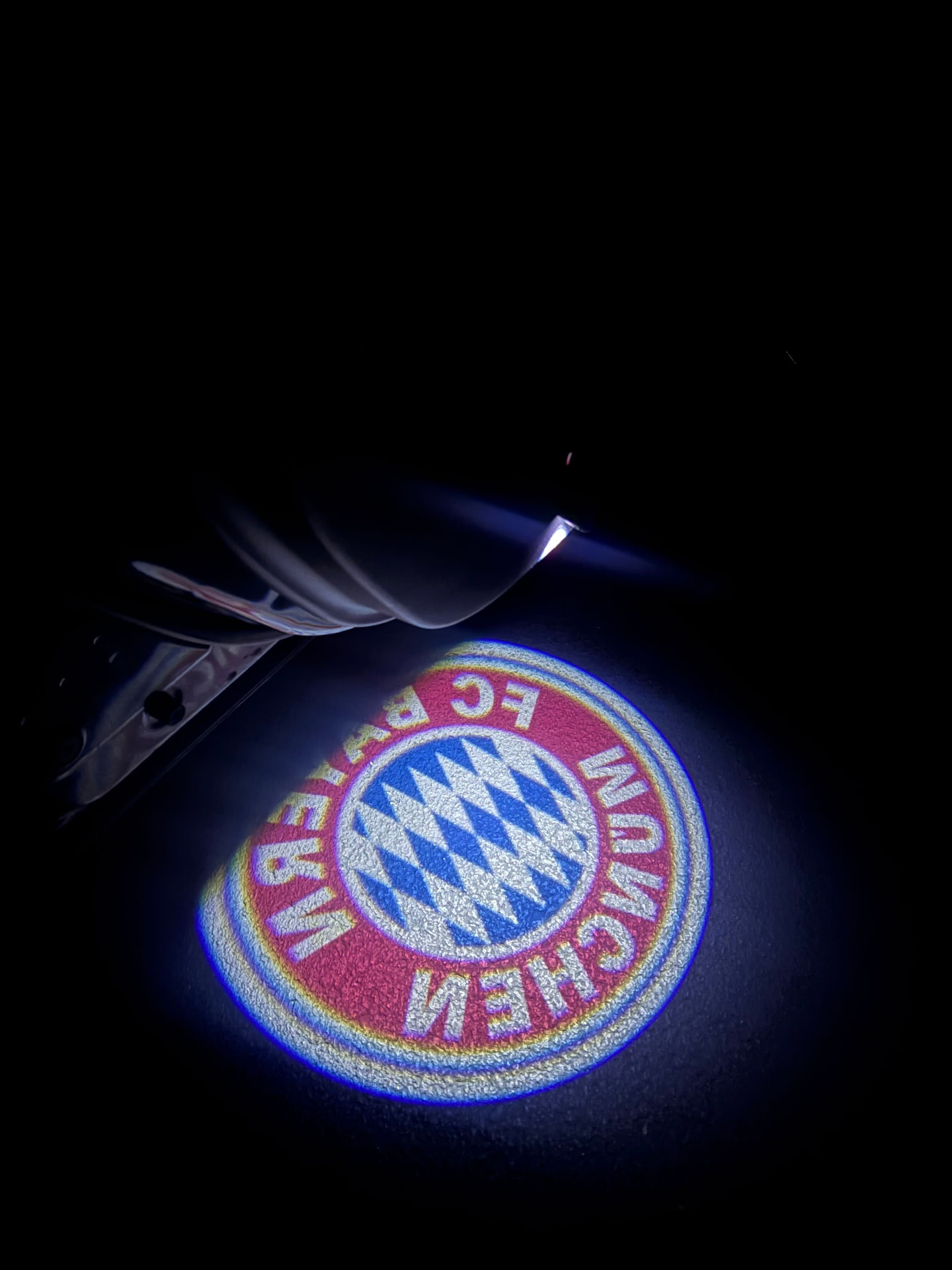 FC Bayern Munich crest projected onto dark asphalt from a DoorBeam unit under an open car door at night